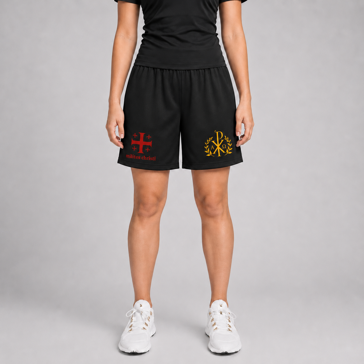 A model from the waist down wearing black athletic shorts with red and gold printed logos, paired with white sneakers, against a neutral gray studio background.