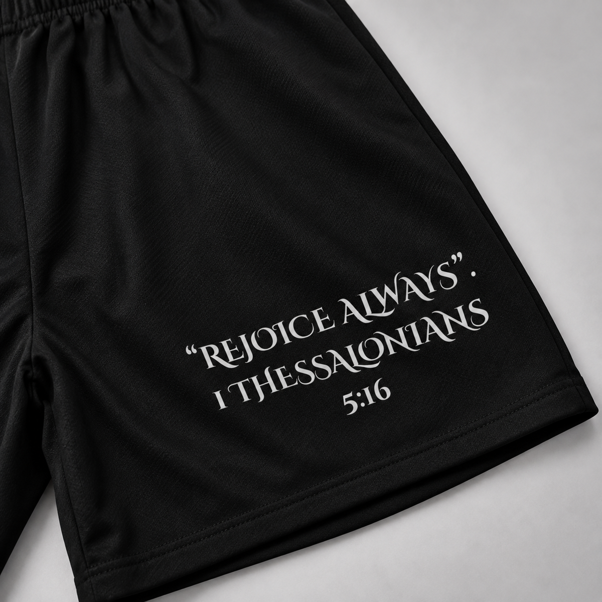 Black athletic shorts with an elastic waistband and white printed text reading “REJOICE ALWAYS. I THESSALONIANS 5:16” on the lower right leg, shown flat against a light gray background.