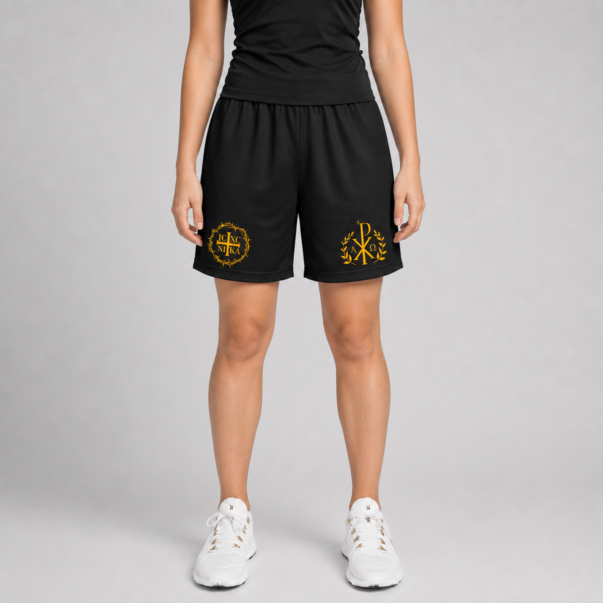 A model is shown from the waist down wearing black athletic shorts with yellow embroidered logos on the left and right legs, paired with a black top and white sneakers, against a light gray studio background.