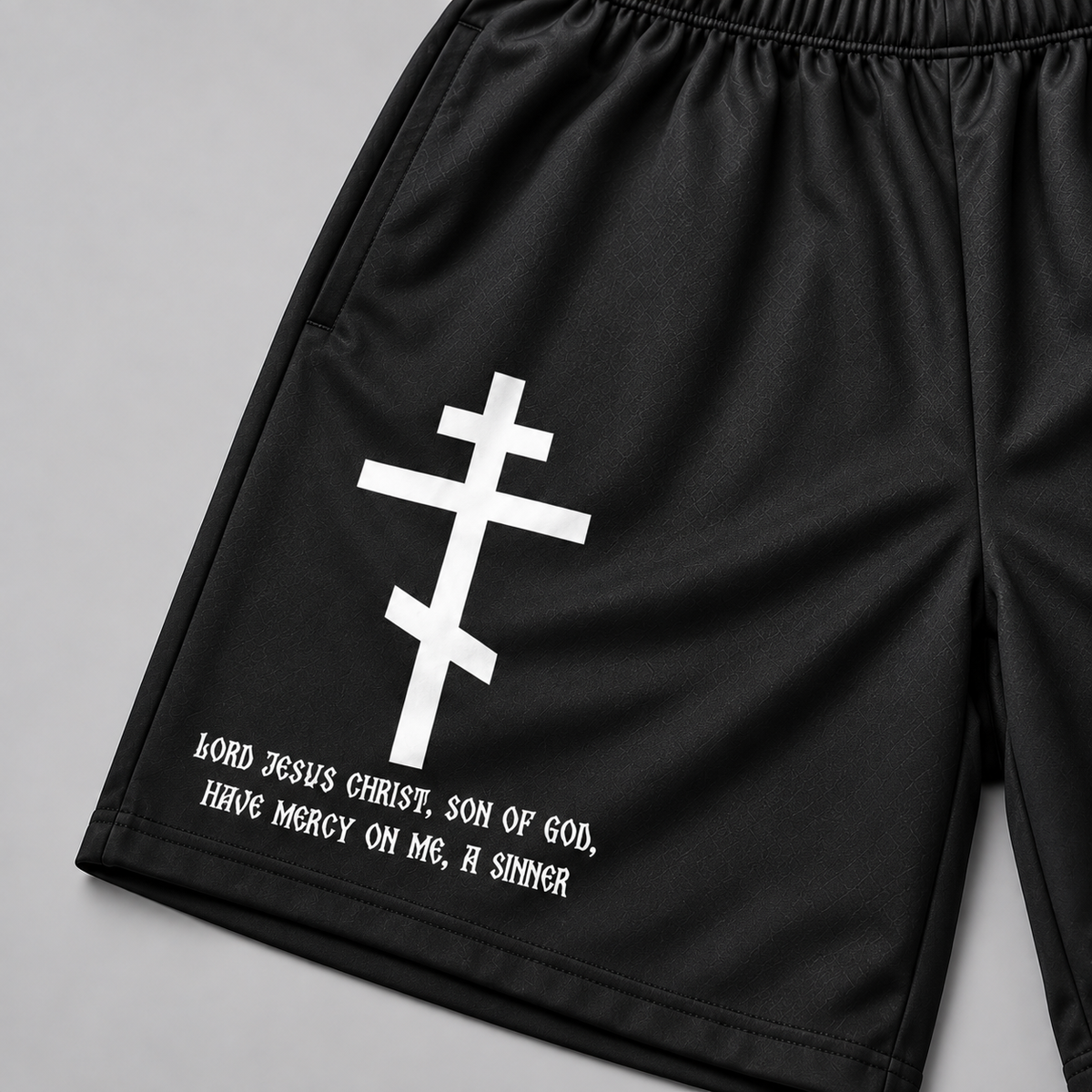 Black athletic shorts with a white Eastern Orthodox cross and the text 'Lord Jesus Christ, Son of God, Have Mercy On Me, A Sinner' printed on the left leg, shown on a neutral gray background.
