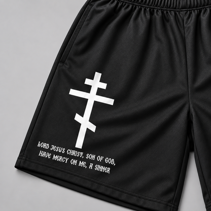 Black athletic shorts with a white Eastern Orthodox cross and the text 'Lord Jesus Christ, Son of God, Have Mercy On Me, A Sinner' printed on the left leg, shown on a neutral gray background.