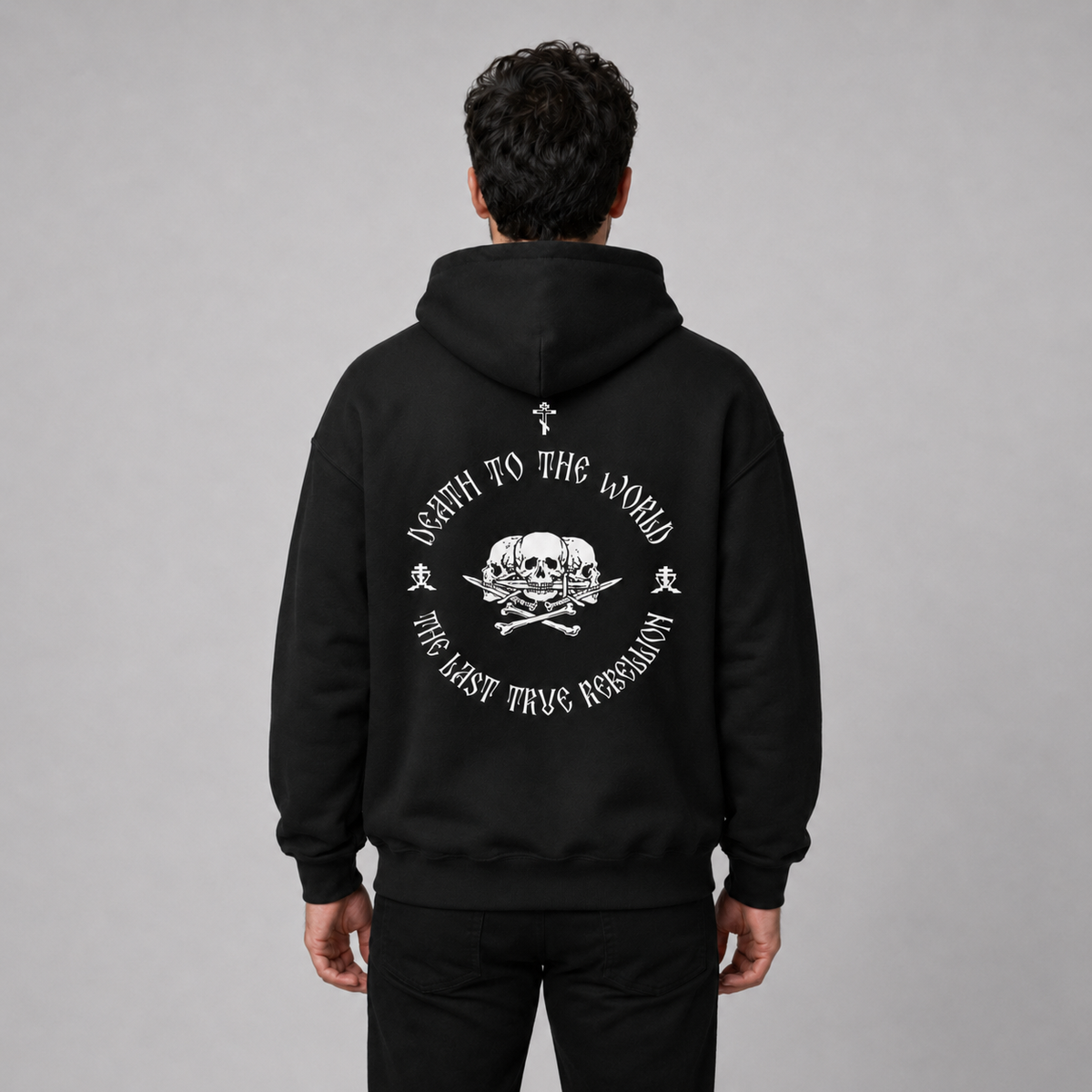 Rückansicht eines Mannes in schwarzem Hoodie mit großem weißem Rückendruck. Das Motiv zeigt einen Totenkopf mit gekreuzten Knochen und kreisförmig angeordneter Aufschrift „THE LAST TRUE REBELLION“.