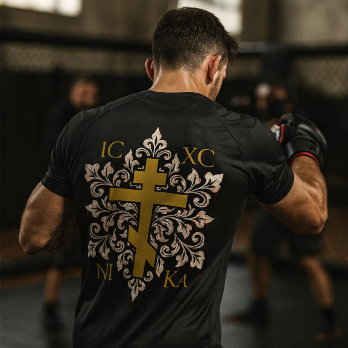Person training in an mma gym wearing a black t-shirt with a gold and white design and text in an indoor setting.