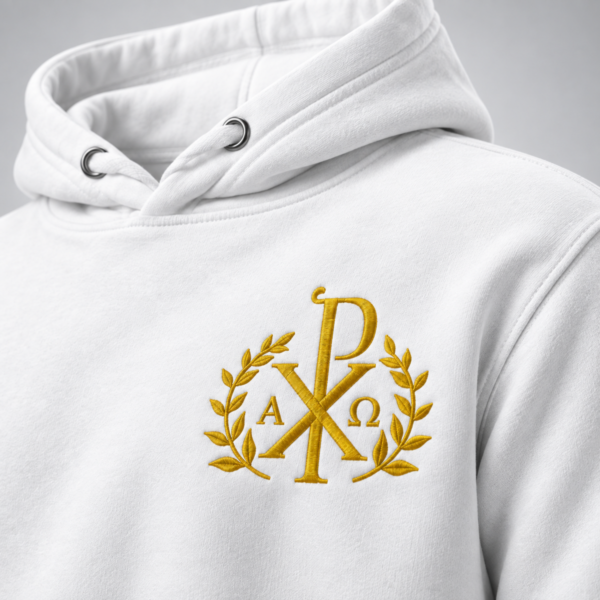 Nahaufnahme eines weißen Hoodies mit goldgesticktem Chi-Rho-Monogramm zwischen Lorbeerzweigen und den griechischen Buchstaben Alpha und Omega darunter vor neutralem grauem Hintergrund.