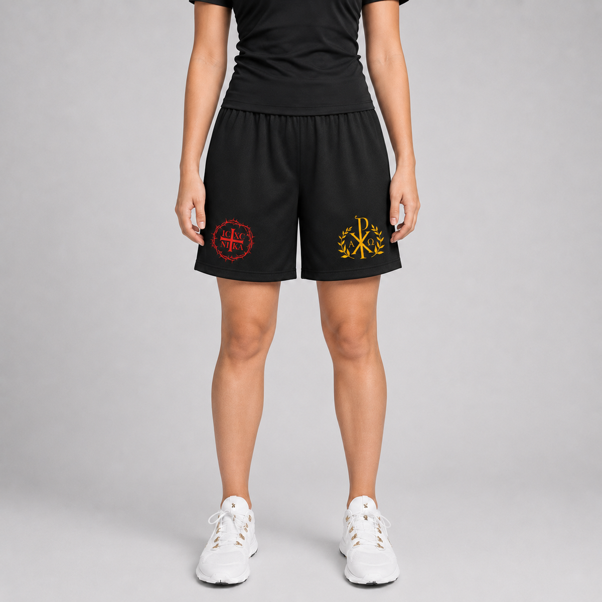 A model is shown from the waist down wearing black athletic shorts with two embroidered logos, one red and one gold, on the left and right legs. The model is also wearing a black t-shirt and white sneakers against a neutral gray studio background.