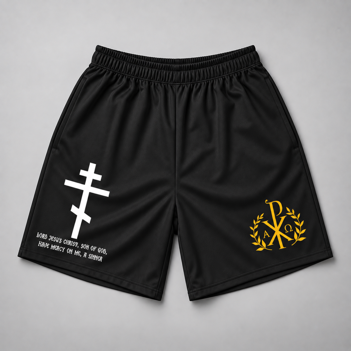 Black athletic shorts with an elastic waistband, featuring a white Orthodox cross with Greek letters on the left leg and a yellow Chi-Rho emblem with laurel leaves on the right leg, displayed flat on a gray background.