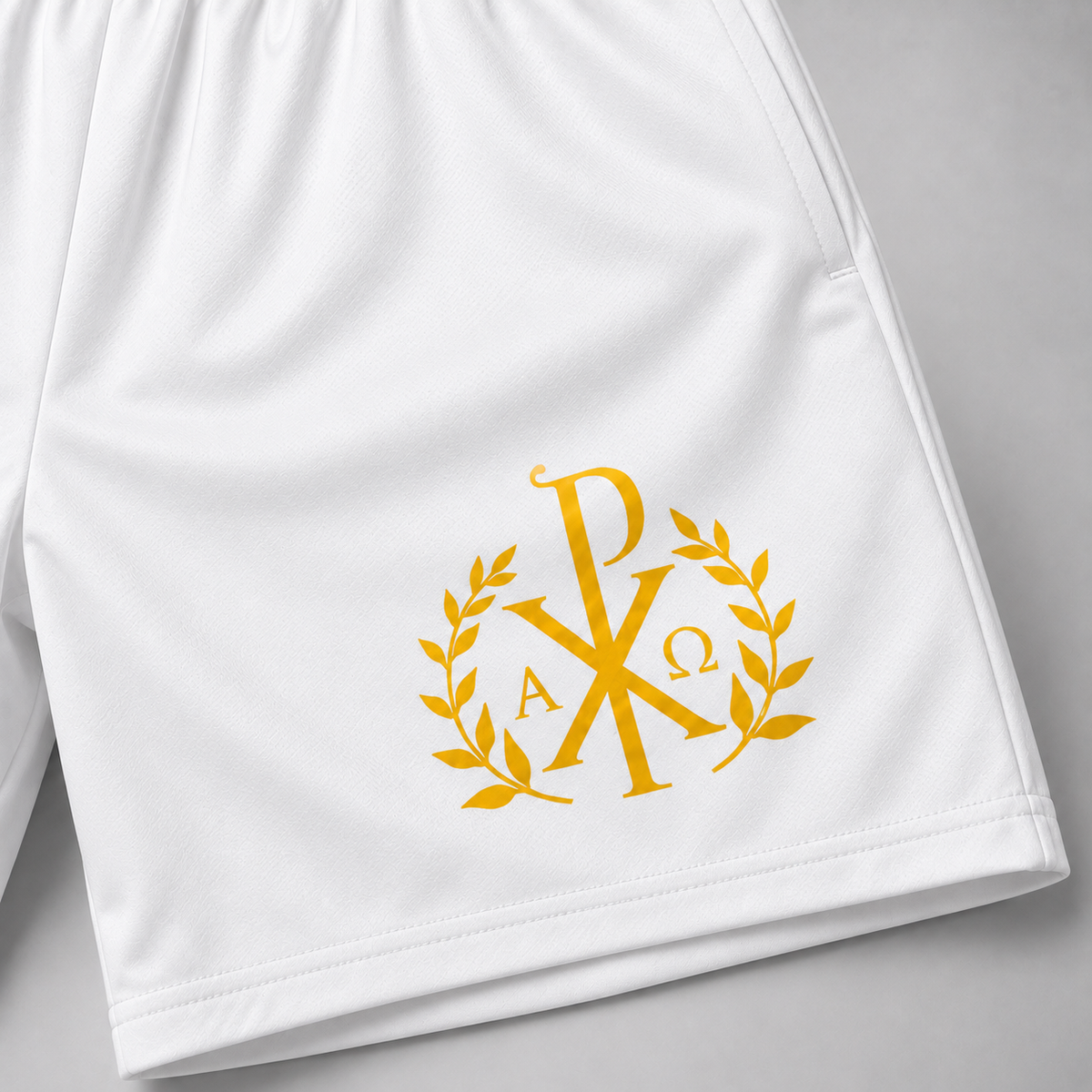 White athletic shorts with a gold monogram logo featuring a Chi-Rho, laurel wreath, and the letters Alpha and Omega, shown on a light gray background.