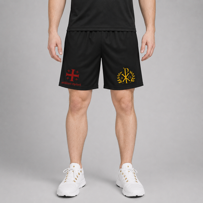 A person wearing black athletic shorts with a red Jerusalem cross and red 'milites christi' text on the left leg, and a gold monogram on the right leg, paired with white sneakers against a gray studio background.