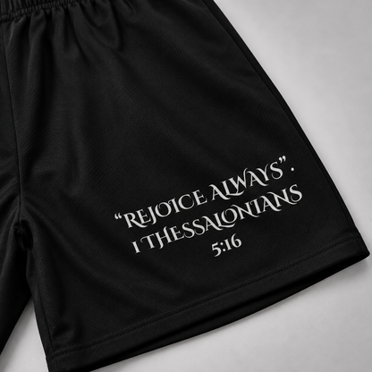 Black athletic shorts with an elastic waistband and white printed text reading “REJOICE ALWAYS. I THESSALONIANS 5:16” on the lower right leg, shown flat against a light gray background.