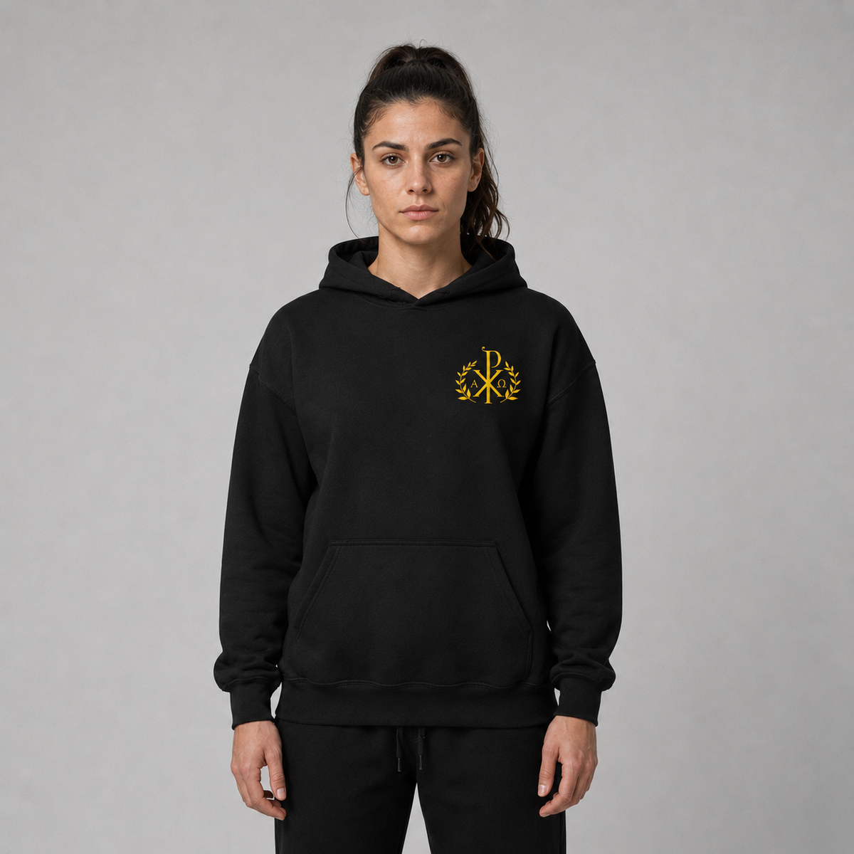Eine Frau präsentiert einen schwarzen Pullover-Hoodie mit kleinem gelb gesticktem Monogramm-Logo links auf der Brust vor schlichtem grauem Studiohintergrund.