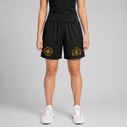 A model is shown from the waist down wearing black athletic shorts with yellow embroidered logos on the left and right legs, paired with a black top and white sneakers, against a light gray studio background.