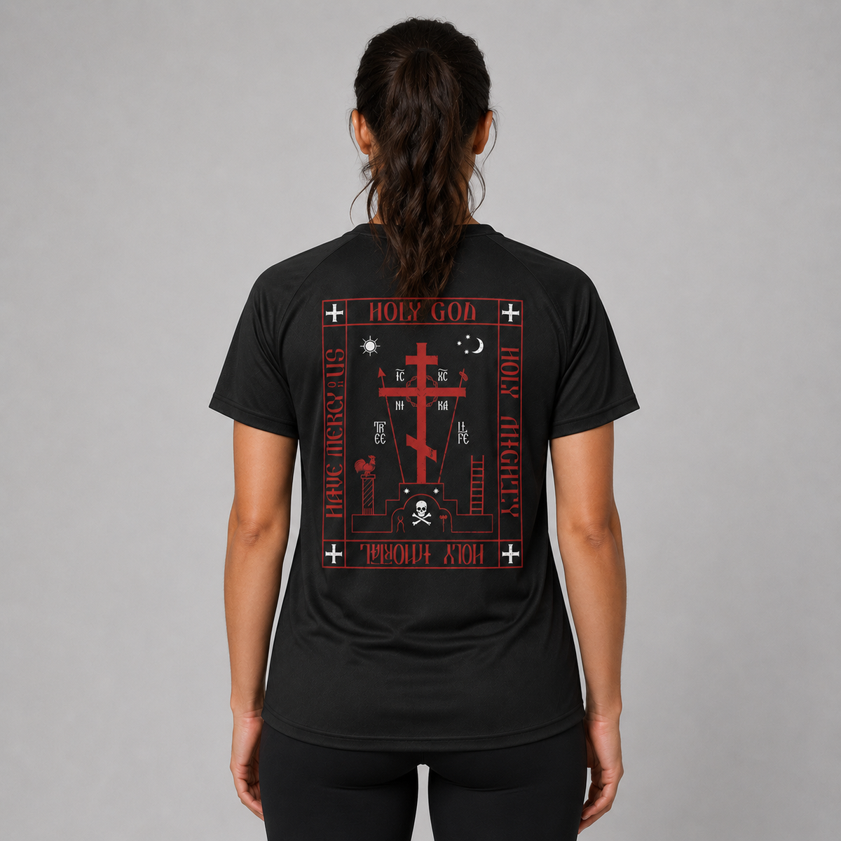 O femeie văzută din spate, purtând un tricou negru cu mâneci scurte și imprimeu mare gotic roșu și alb, împreună cu colanți negri, pe un fundal gri neutru de studio.