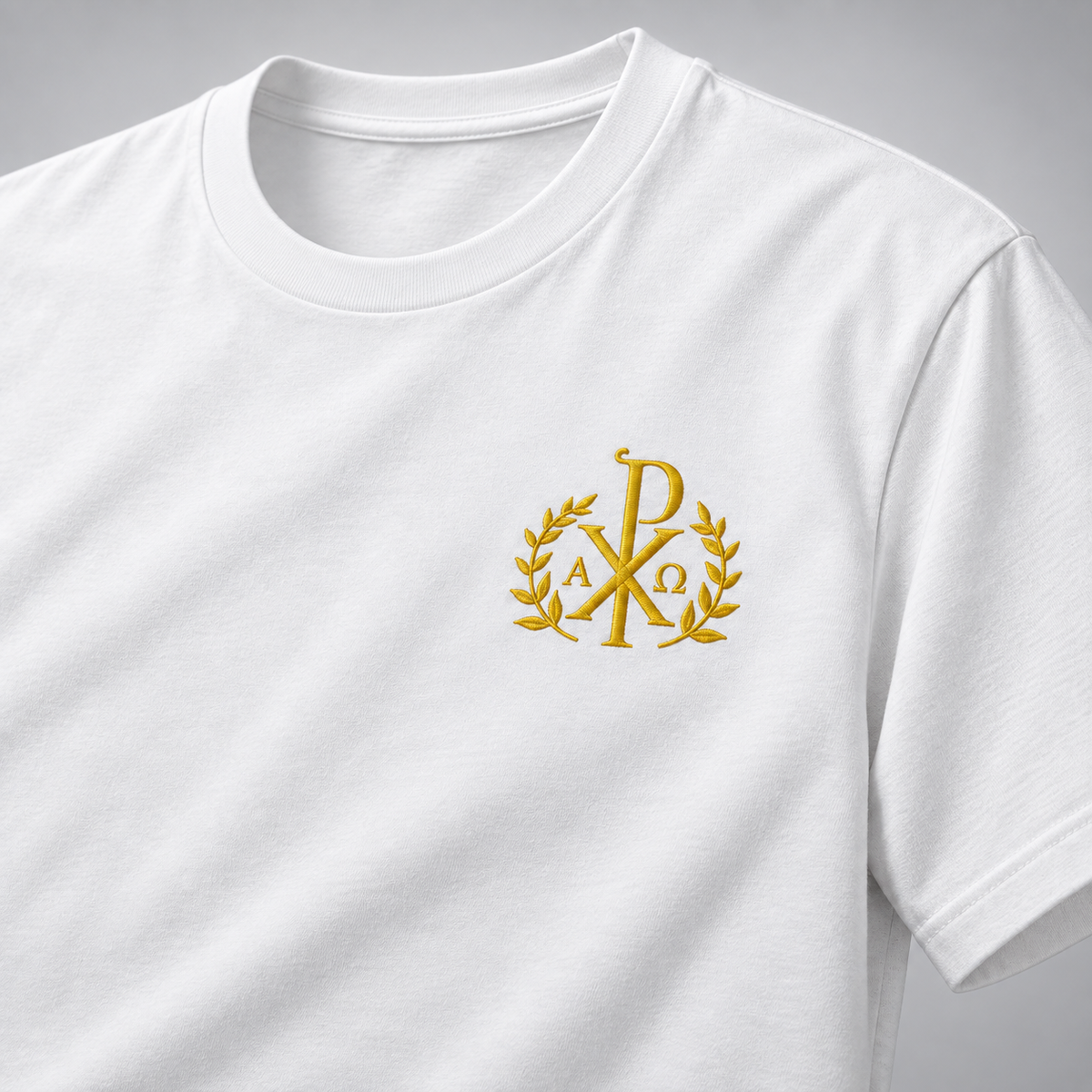 Detaliu al unui tricou alb cu mâneci scurte, decolteu rotund și monogramă aurie brodată în stânga pieptului, pe un fundal gri neutru.