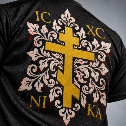 Close-up of the back of a black mesh athletic jersey featuring a large gold cross over an ornate cream damask-style motif with red dripping accents and gold text reading IC XC NI KA.