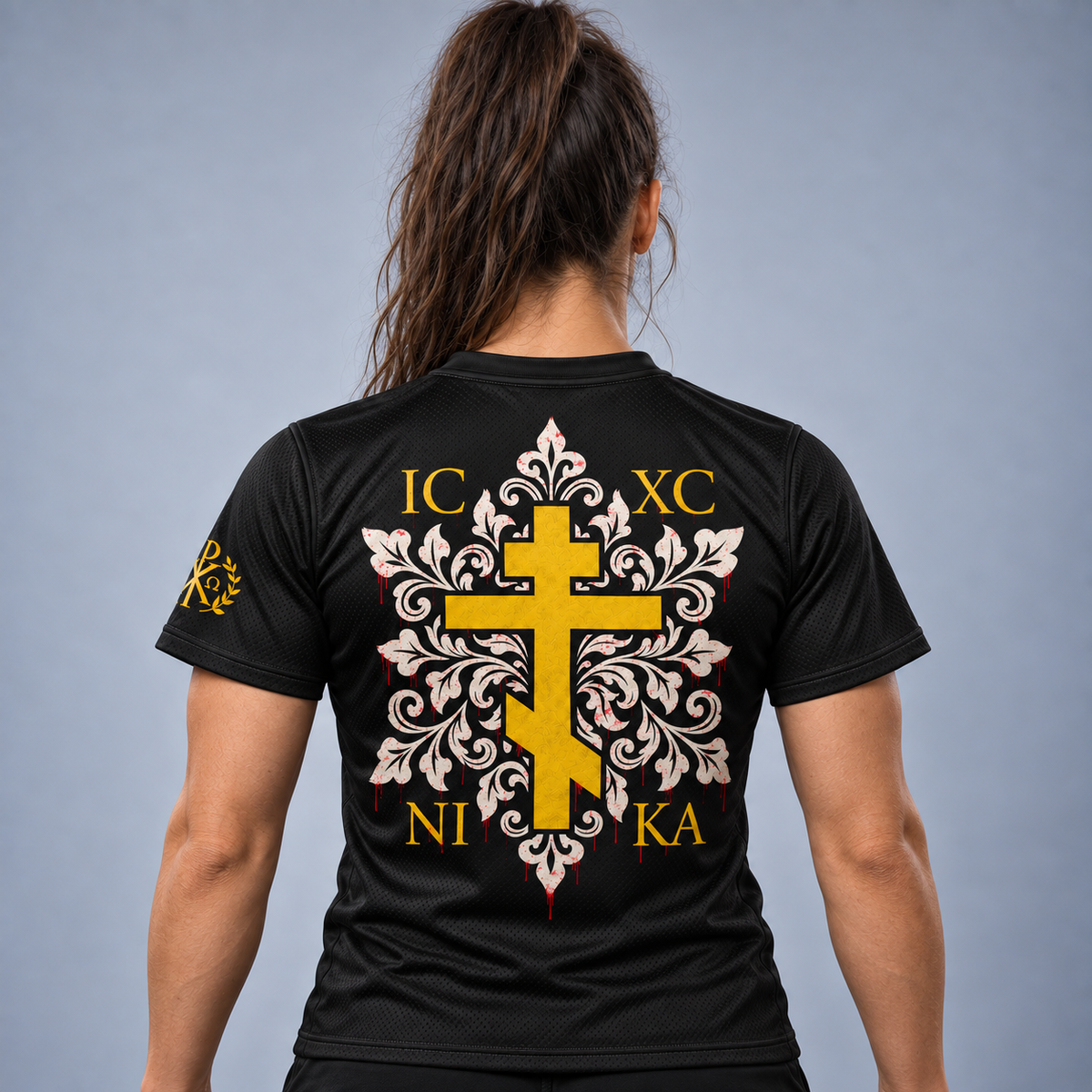 Rear view of a woman wearing a black short-sleeve athletic T-shirt with a large yellow and white ornamental cross graphic and gold lettering on the back, against a light gray studio background.