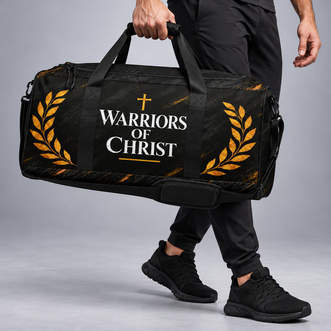 A man in a black t-shirt and black athletic pants carries a black duffel bag with gold laurel wreaths and the text 'WARRIORS OF CHRIST' printed on the front, set against a gray studio background.