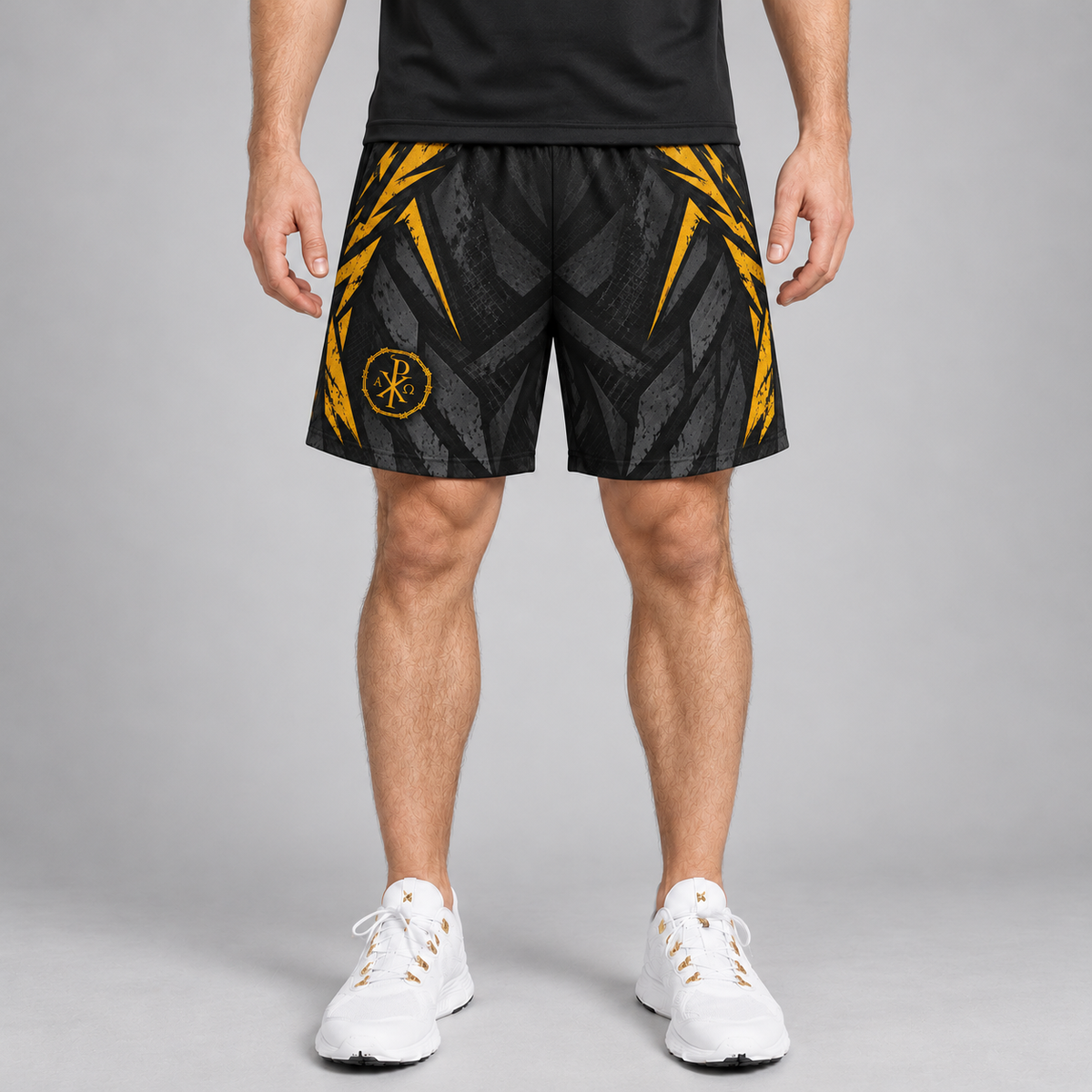 A man wearing black athletic shorts with a yellow and gray geometric pattern and a circular logo on the left leg, paired with a black shirt and white sneakers, standing against a gray studio background.