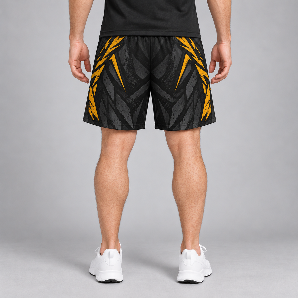 A person shown from the back wearing black athletic shorts with a bold yellow geometric pattern, a black shirt, and white sneakers against a gray studio background.