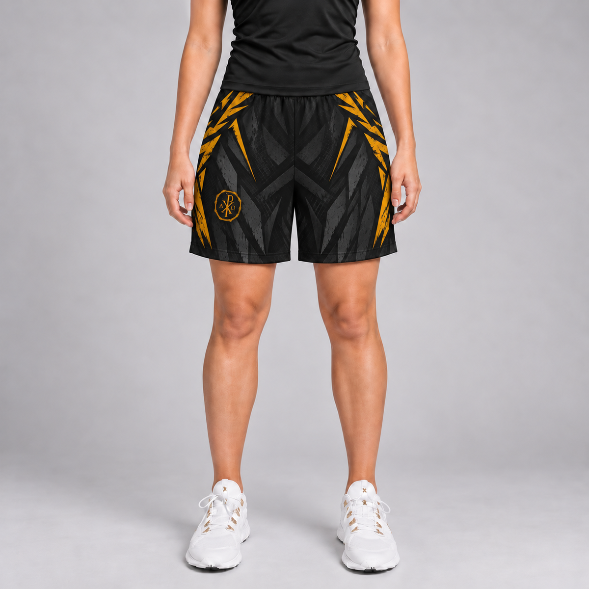 A model from the waist down wearing black athletic shorts with a gold geometric pattern and a circular logo on the left leg, paired with a black top and white sneakers, against a gray studio background.