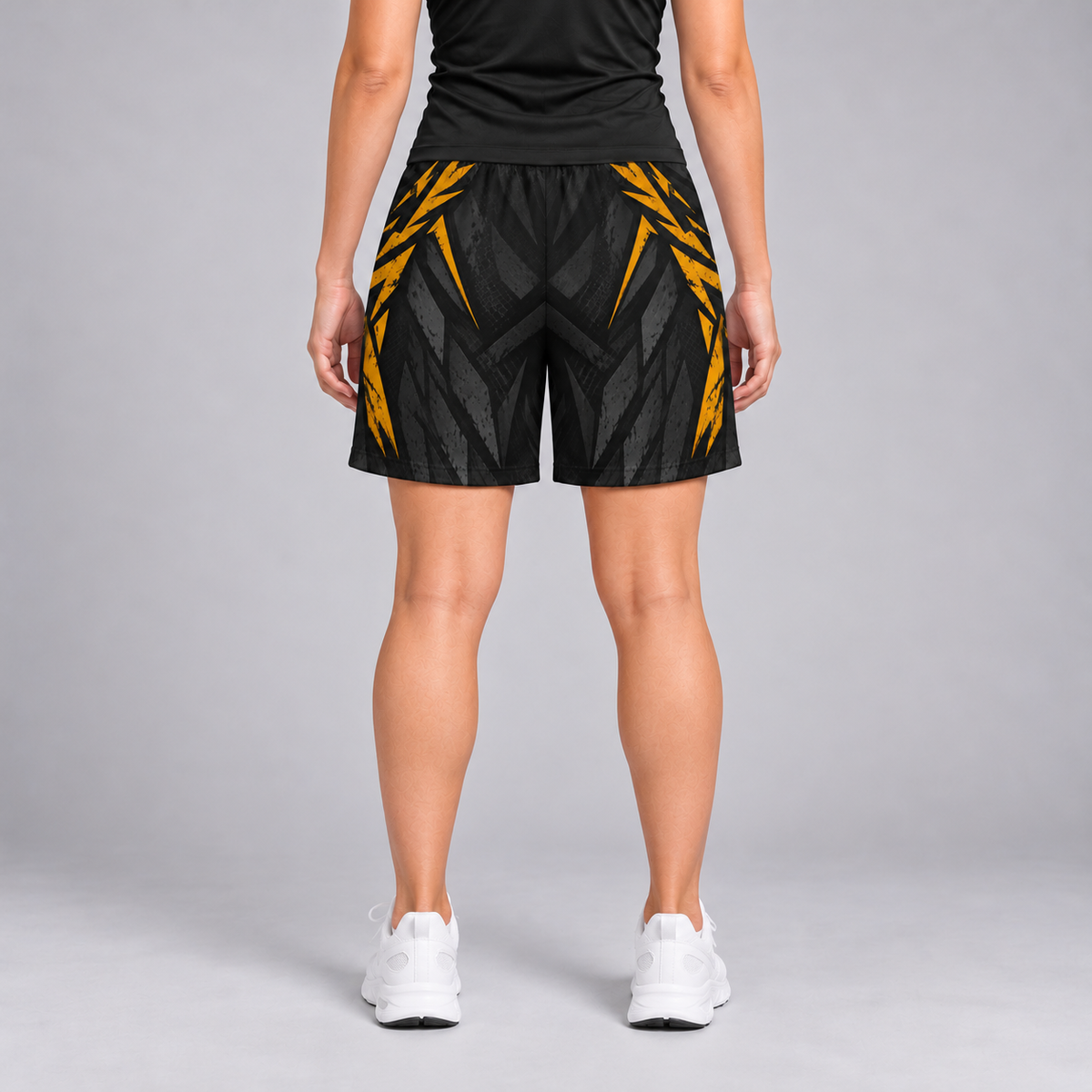 A person shown from the back wearing black athletic shorts with a bold yellow geometric pattern, a black athletic top, and white sneakers against a light gray studio background.