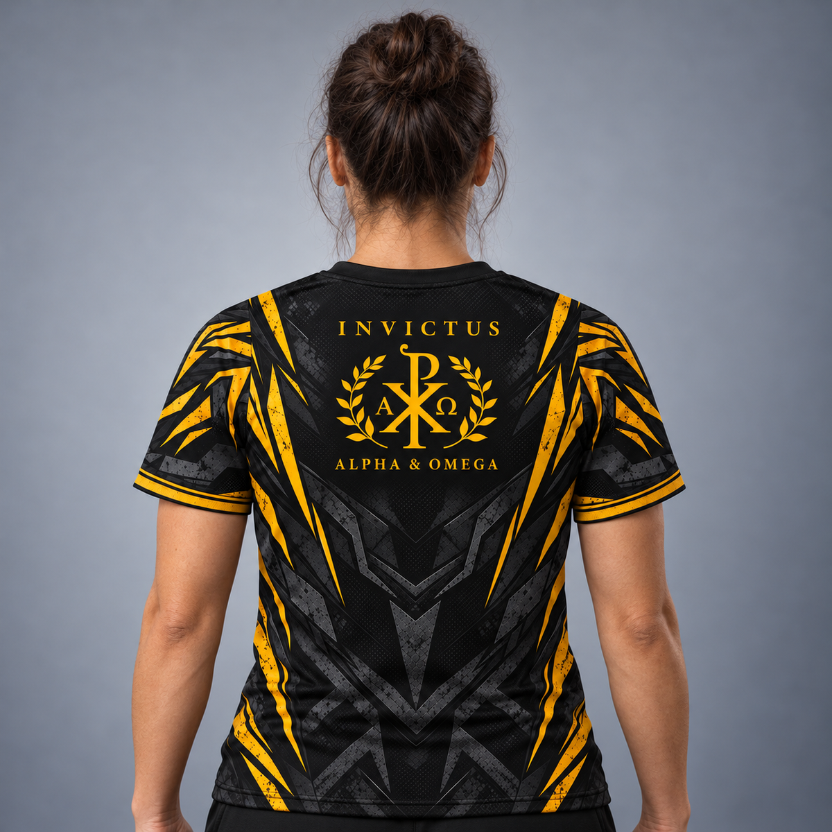 A woman viewed from behind wearing a black short-sleeve athletic jersey with bold yellow and gray graphic patterns and a large 'INVICTUS ALPHA & OMEGA' crest on the back, against a plain gray studio background.