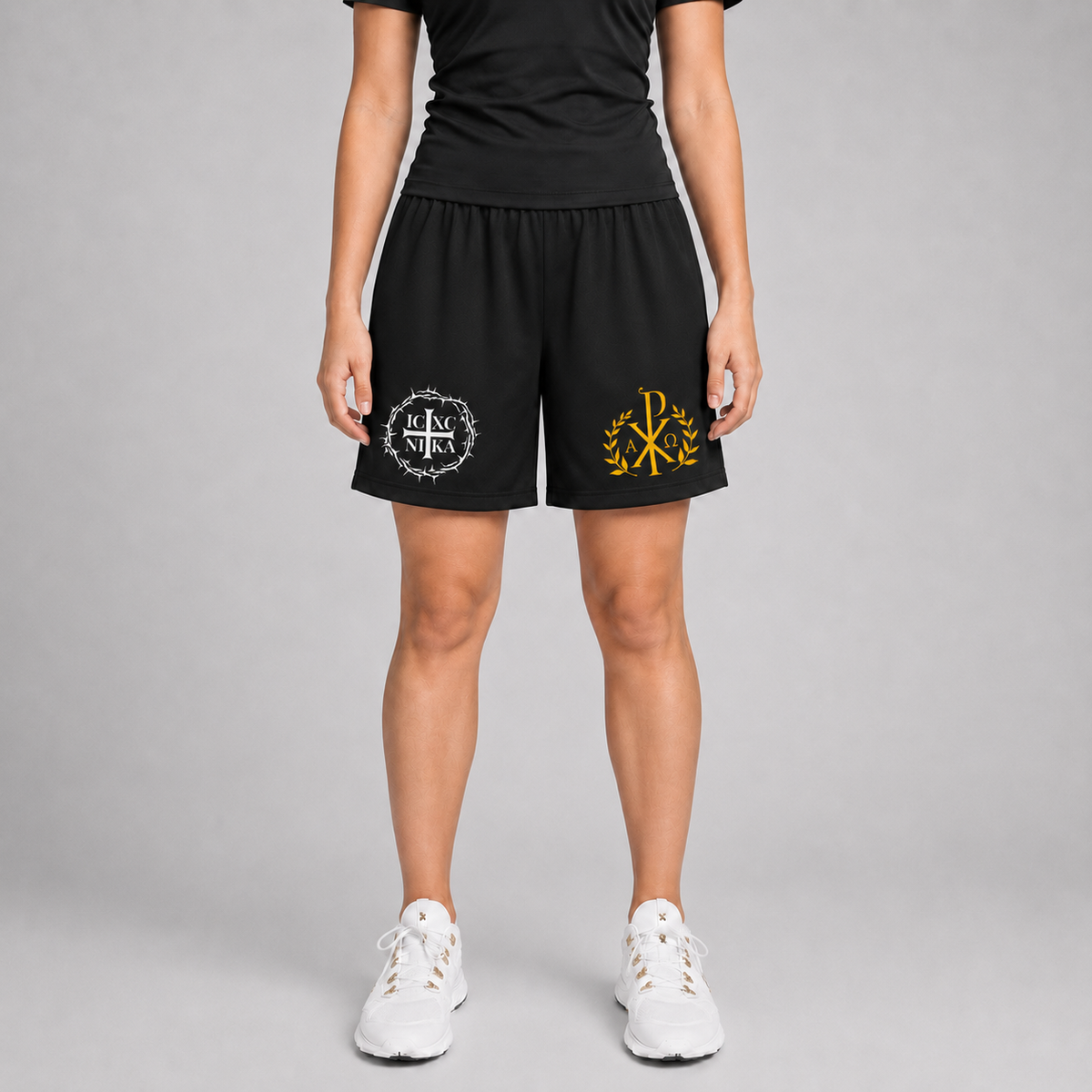 A model from the waist down wearing black athletic shorts with a white circular monogram logo on the left leg and a yellow Chi-Rho monogram logo on the right leg, paired with white sneakers against a gray studio background.
