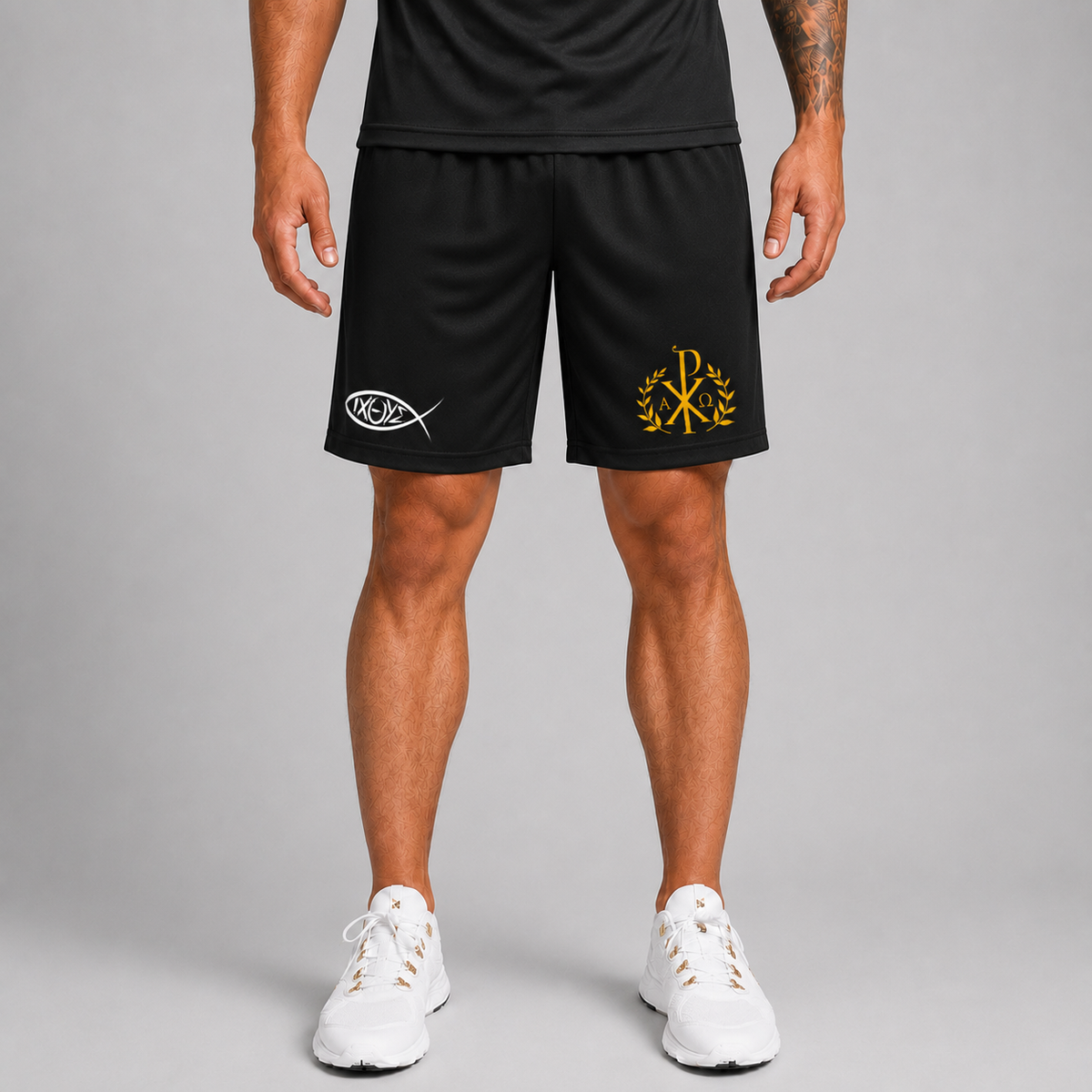 Un bărbat văzut de la talie în jos, purtând pantaloni scurți sport negri cu pește alb pe piciorul stâng și monogramă aurie pe cel drept, cu pantofi sport albi, pe un fundal gri de studio.