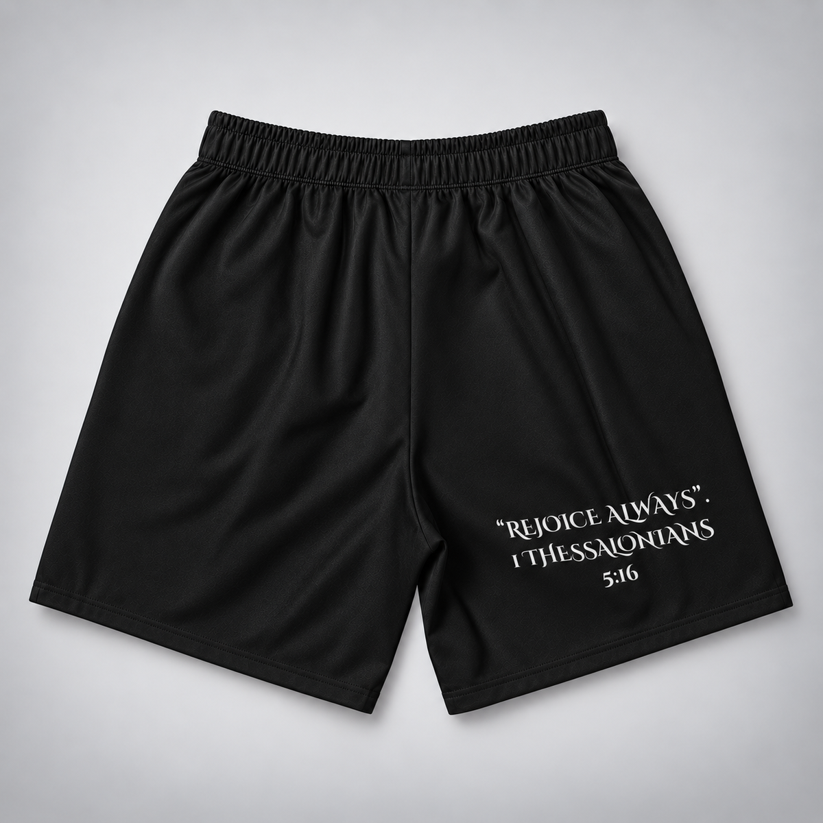 Black athletic shorts with an elastic waistband and white printed text reading “REJOICE ALWAYS. I THESSALONIANS 5:16” on the lower right leg, shown flat against a light gray background.