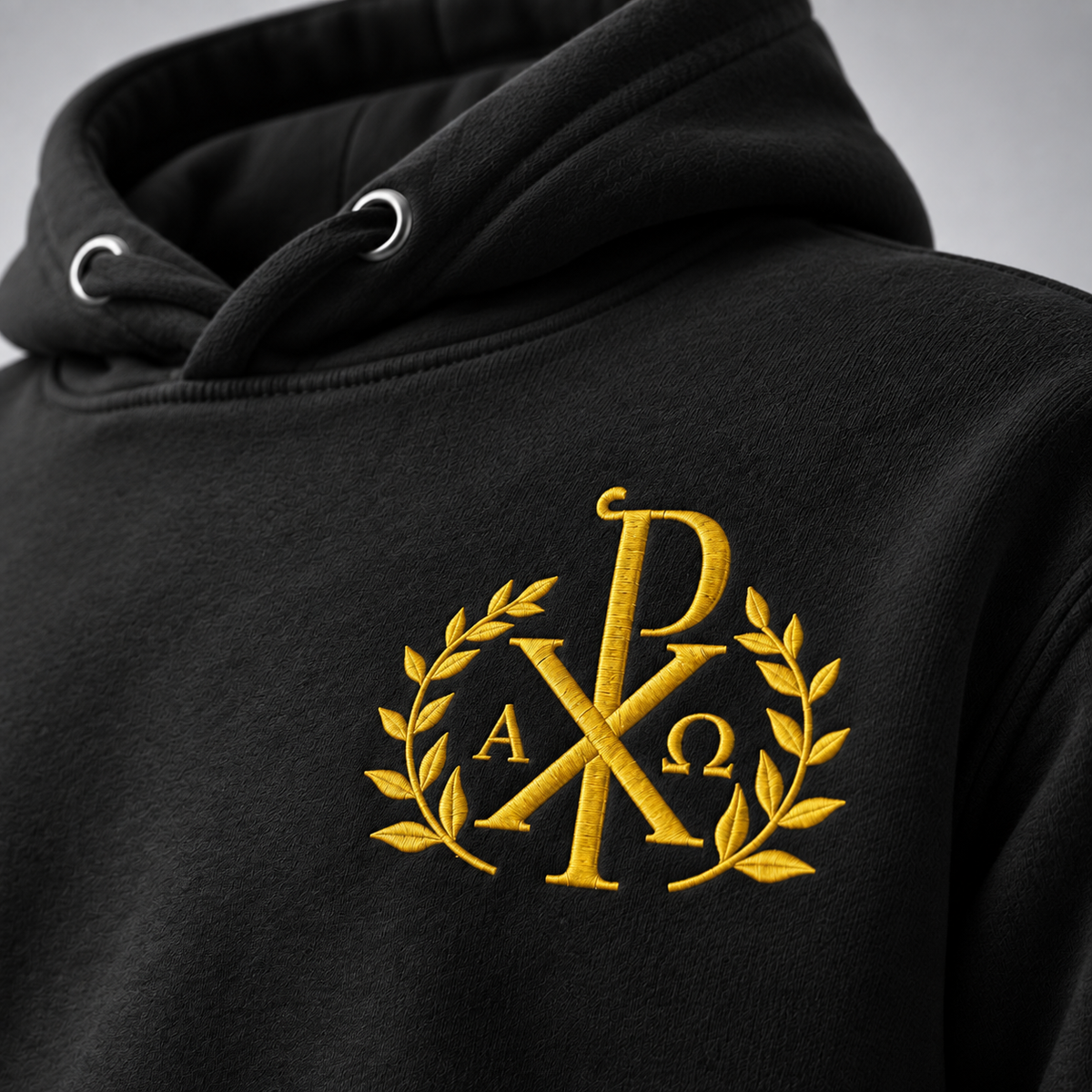 Close-up of a black hoodie with a gold embroidered monogram logo on the chest, featuring the letters Alpha and Omega enclosed in a laurel wreath.
