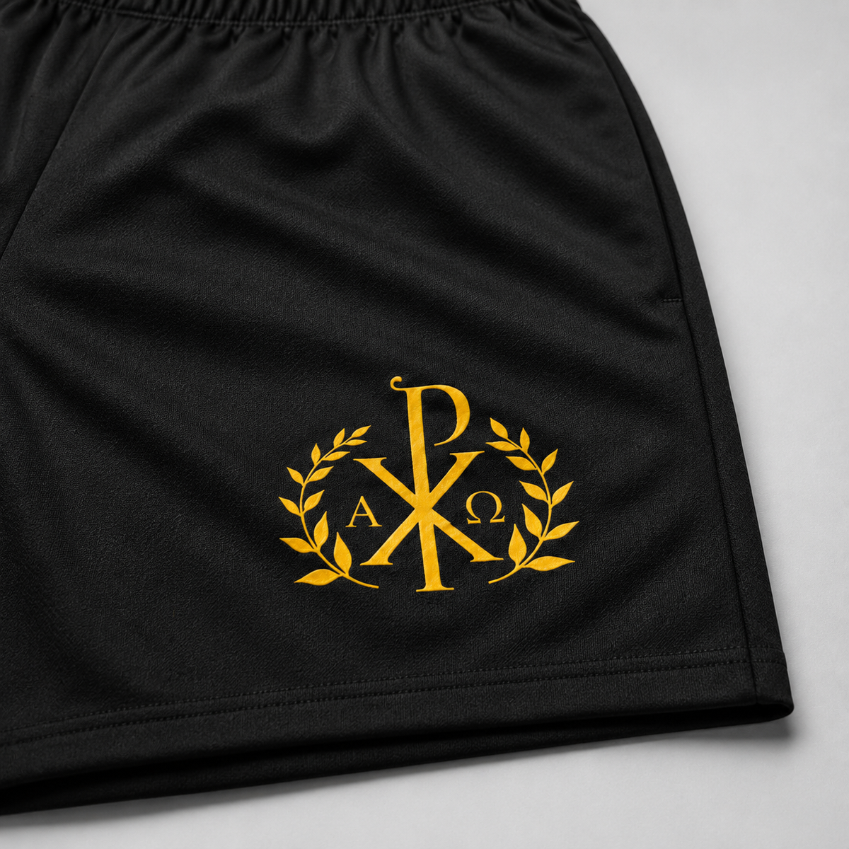 Close-up of black athletic shorts with a gold embroidered monogram logo featuring the Chi-Rho, laurel leaves, and the letters Alpha and Omega, set against a light gray background.