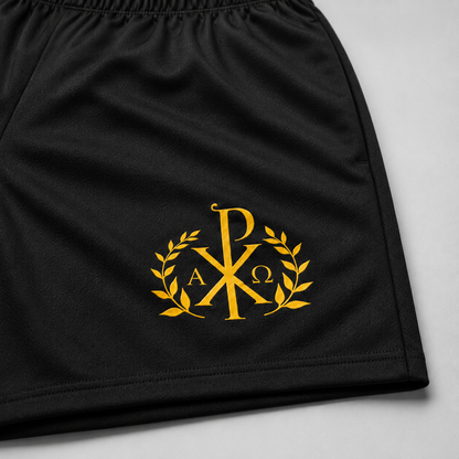Detaliu al unor pantaloni scurți sport negri, cu monogramă aurie brodată reprezentând Hristograma, frunze de laur și literele Alfa și Omega, pe un fundal gri deschis.