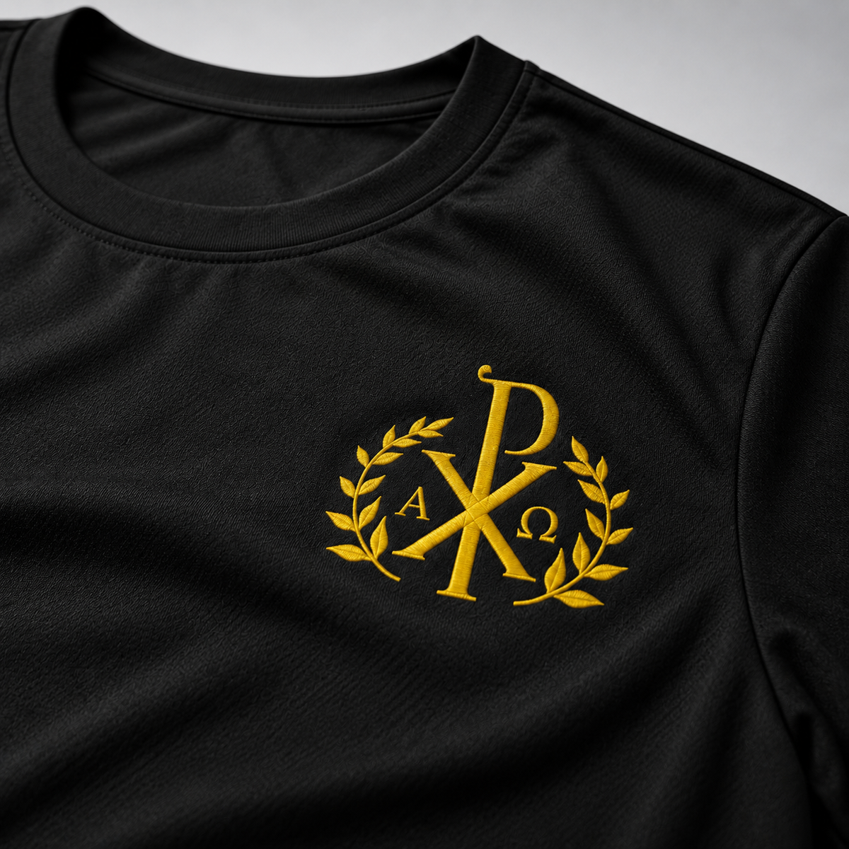 Nahaufnahme eines schwarzen Rundhals-T-Shirts mit goldgesticktem Chi-Rho-Monogramm, Lorbeerzweigen und den griechischen Buchstaben Alpha und Omega auf der Brust.