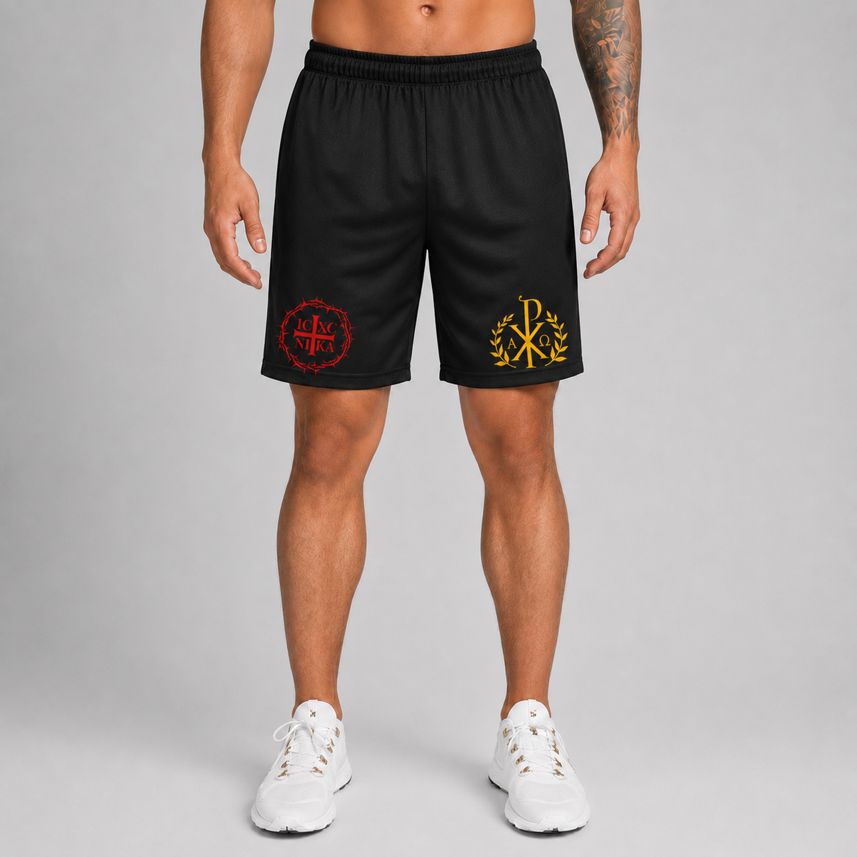A man modeling black athletic shorts with red and yellow embroidered logos, paired with white sneakers, against a neutral gray studio background.